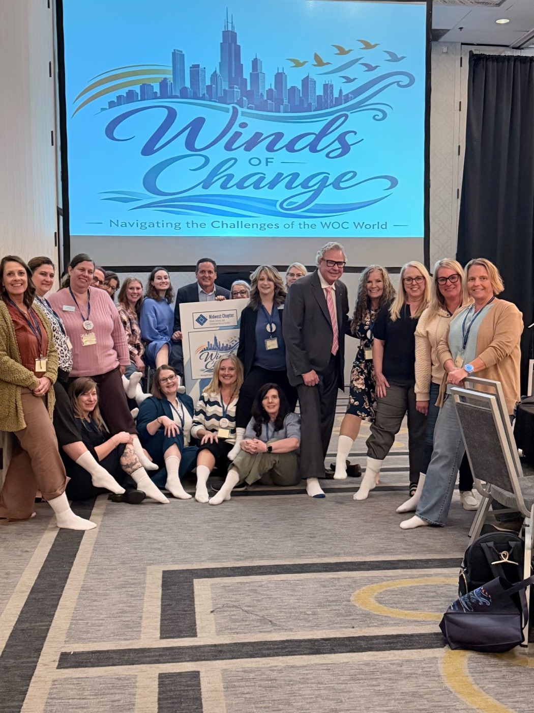 Conference attendees in front of a Winds of Change screen, each wearing a single white sock